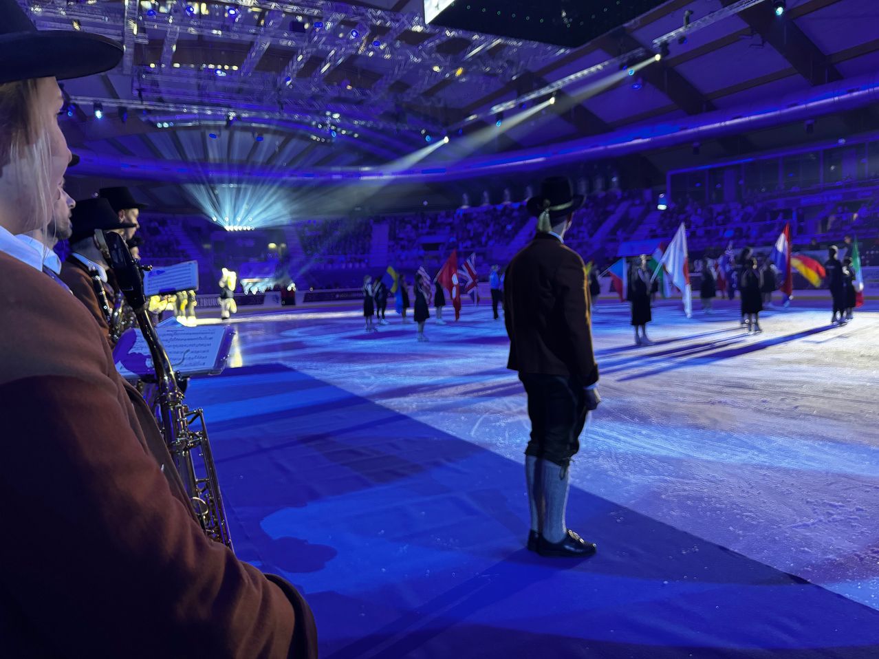 An ice skating event with performers on the ice and flags of various countries. A man stands holding a saxophone. The venue is illuminated with blue lights.