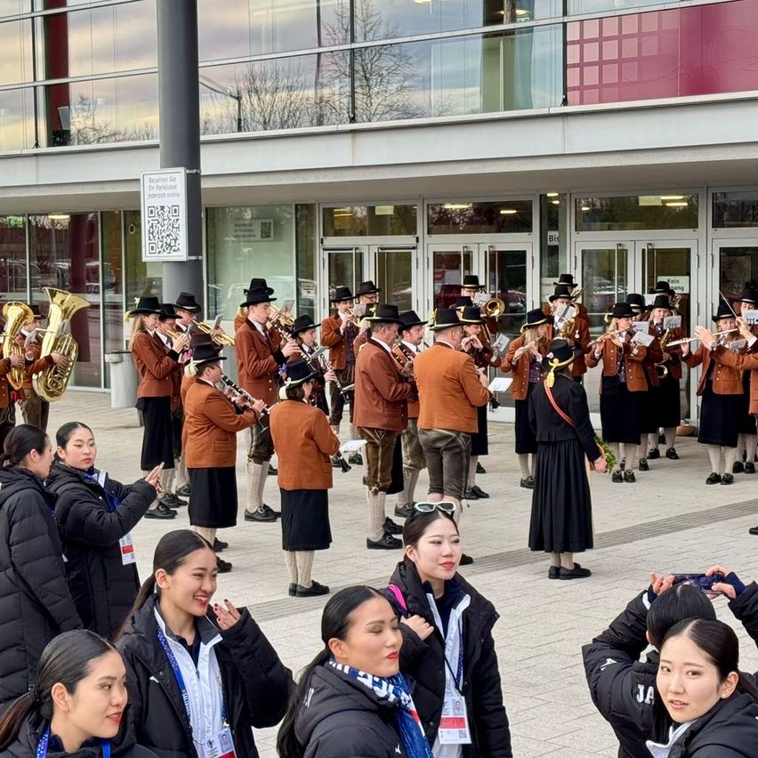 A marching band performs outside a modern building. The musicians, dressed in brown uniforms, play various instruments. Spectators, including women with ID badges, stand nearby and watch.