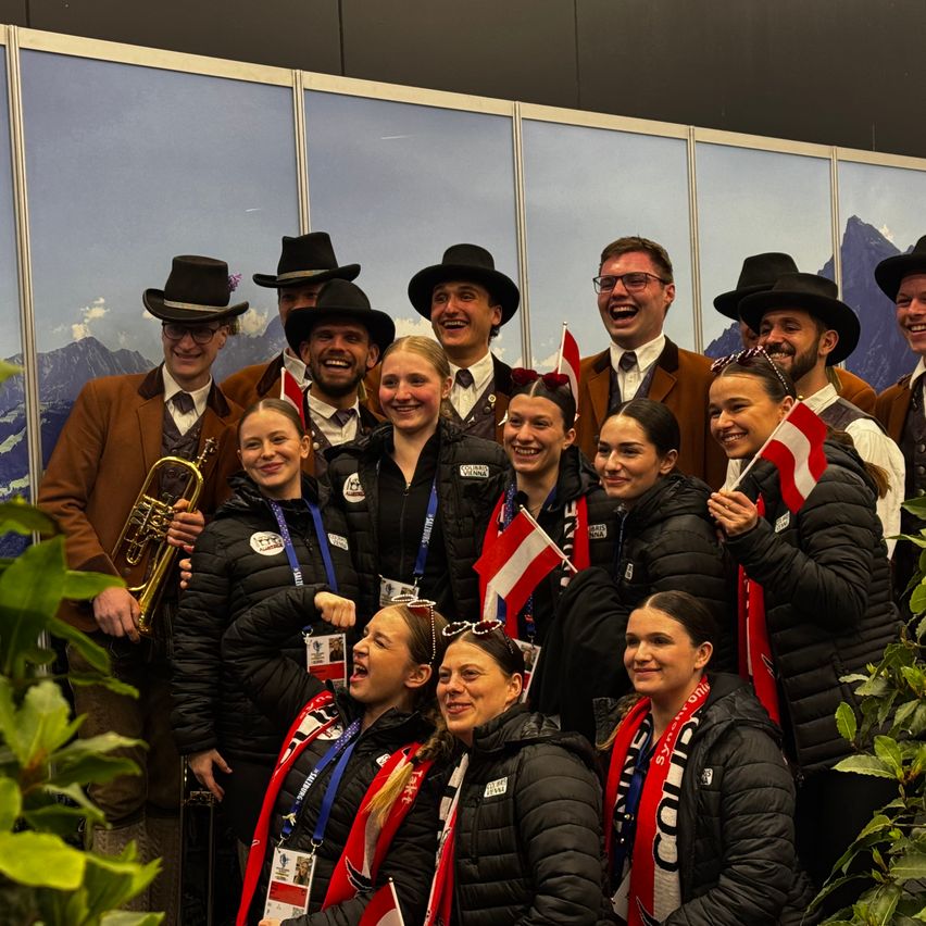 A group of people dressed in traditional costumes, some with hats and others holding flags, are smiling and posing for a photo. They are standing in front of a backdrop with a mountain scene.