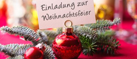 Ein roter Weihnachtsschmuck mit einem Zettel oben drauf, auf dem 'Einladung zur Weihnachtsfeier' steht, liegt auf einem Tisch mit Tannennadeln und einem weiteren roten Ornament.