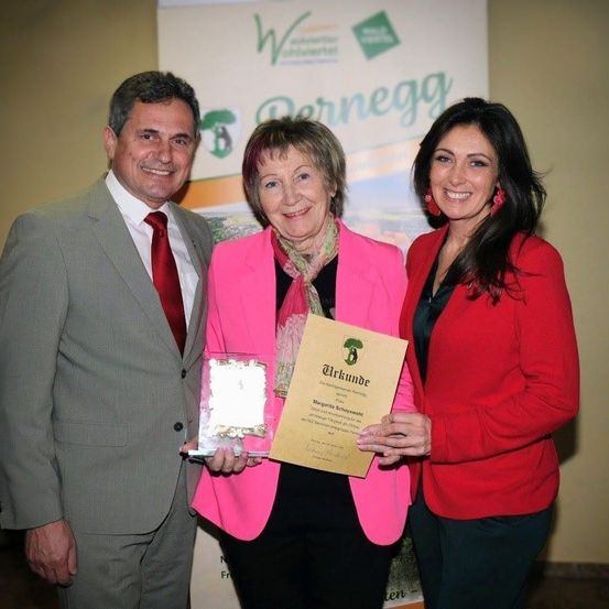 Eine Frau hält eine Auszeichnung in der Hand, während sie zwischen einem Mann und einer weiteren Frau steht, alle lächeln für ein Foto. Hinter ihnen ist ein Banner mit dem Wort 'Bernegg'.