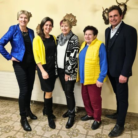 Fünf Personen stehen in einer Reihe, lächelnd, vor einer Wand mit Hirschgeweihen und einem Heizkörper. Sie tragen verschiedene Farben und Stile von Kleidung.