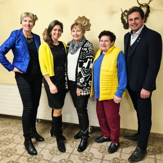 Fünf Personen stehen in einer Reihe, lächelnd, vor einer Wand mit Hirschgeweihen und einem Heizkörper. Sie tragen verschiedene Farben und Stile von Kleidung.