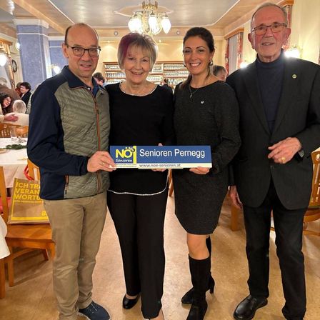 Vier Menschen stehen in einem Restaurant zusammen. Sie halten ein blaues Schild mit weißem Text, auf dem NO! Senioren Pernegg steht. Eine Frau hält das Schild in der Mitte.