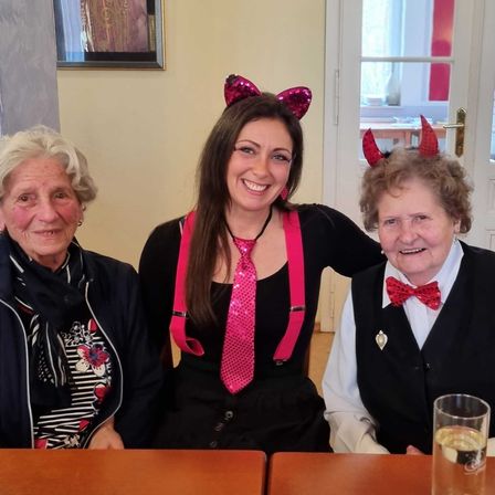 Drei Frauen sitzen gemeinsam an einem Tisch. Eine trägt Teufelshörner, eine andere ein rosafarbenes Halstuch und alle lächeln für ein Foto.