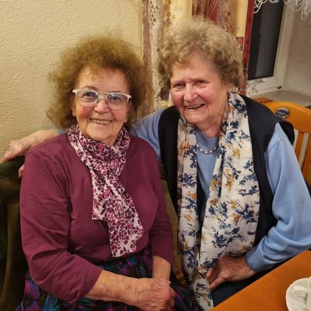 Zwei ältere Frauen sitzen zusammen und lächeln, eine trägt eine Brille und einen Schal, die andere einen Blumenschal und eine Halskette. Sie scheinen sich drinnen zu befinden, in der Nähe eines Fensters mit Vorhängen.