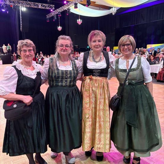 Vier Frauen stehen eng beieinander und lächeln für ein Foto. Sie sind in traditioneller Kleidung gekleidet und scheinen sich auf einem Festival zu befinden. Hinter ihnen befindet sich eine Menschenmenge, die auf Stühlen sitzt und eine Bühne beobachtet.