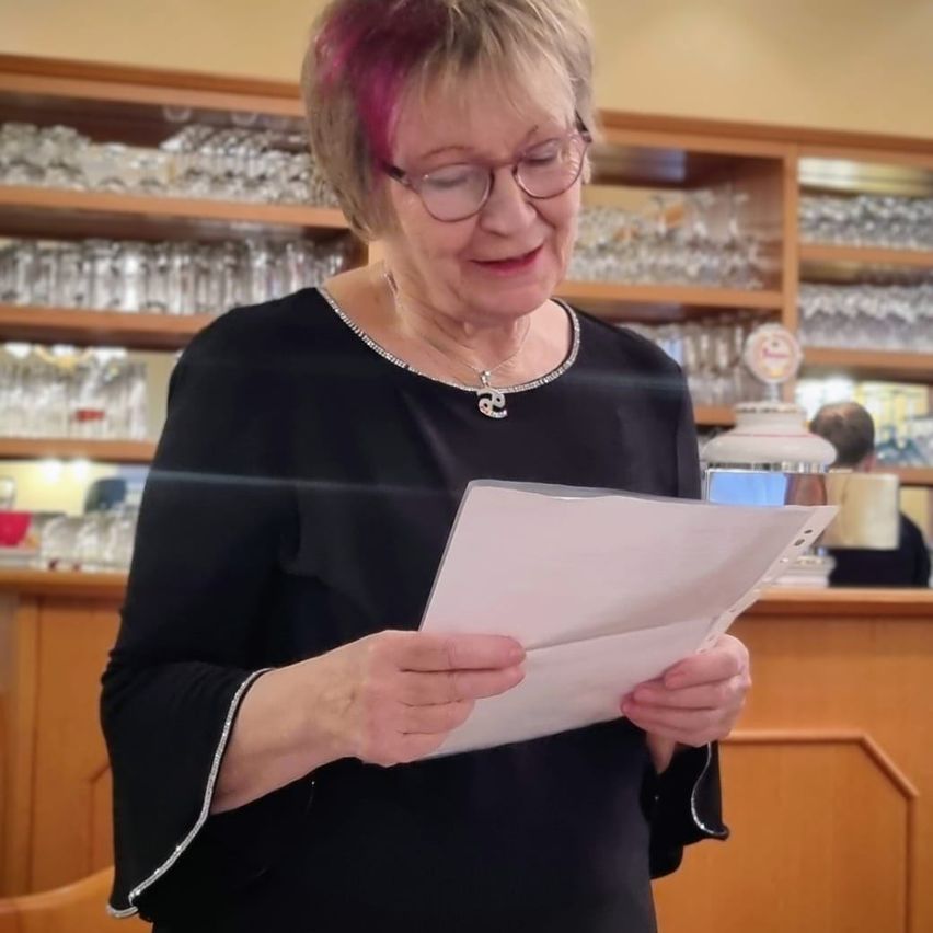 Eine ältere Frau mit pinkem Haar, Brille und Halskette steht, ein Papier in der Hand, in einer Bar mit Regalen voller Gläser im Hintergrund.