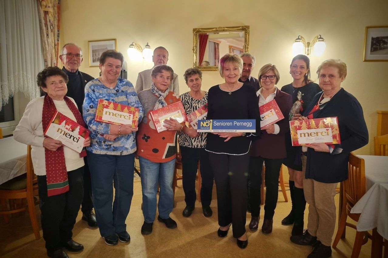 Eine Gruppe älterer Menschen steht lächelnd zusammen und hält Schilder. Eine Person in der Mitte hält ein blaues Schild mit weißen Text. Hinter ihnen spiegelt ein Spiegel einen Raum mit einem goldenen Rahmen.