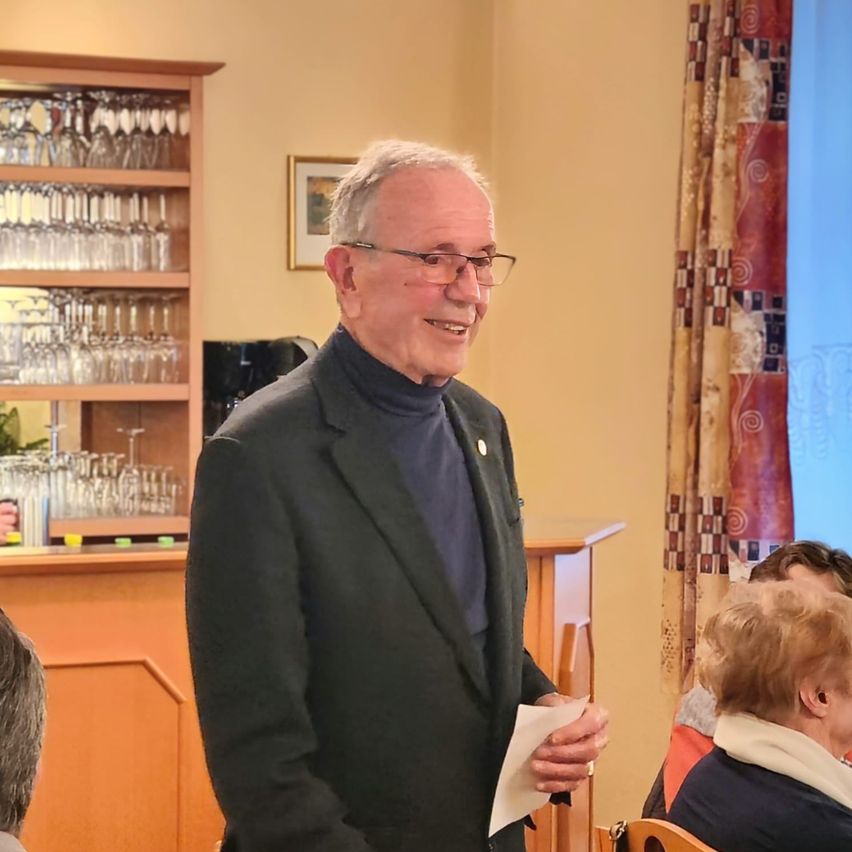 Ein älterer Mann steht und lächelt, während er ein Stück Papier in einem Raum mit einer hölzernen Bar und Regalen voller Gläser hält. Hinter ihm sitzen eine Frau und ein Mann.