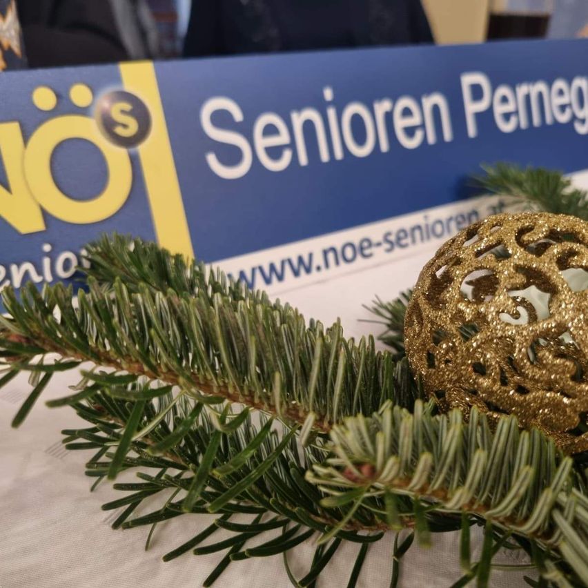 Eine goldene Zierkugel liegt auf einem Tannenzweig. Dahinter befindet sich ein blaues Schild mit den Worten 'Senioren Permeg'. Unter den Zweigen befindet sich eine weiße Tischdecke.