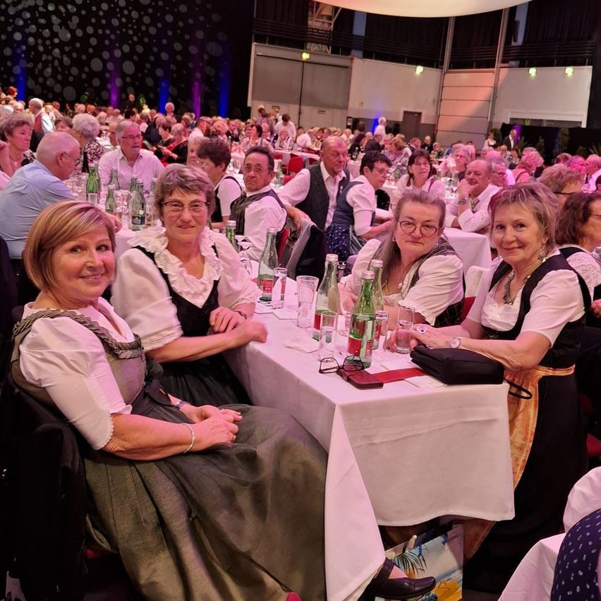 Several adults, mostly women, are seated at a banquet hall. They are dressed in traditional attire and smiling. The table has bottles, glasses, and a wallet. The hall has a stage, lights, and a ceiling with white spots.