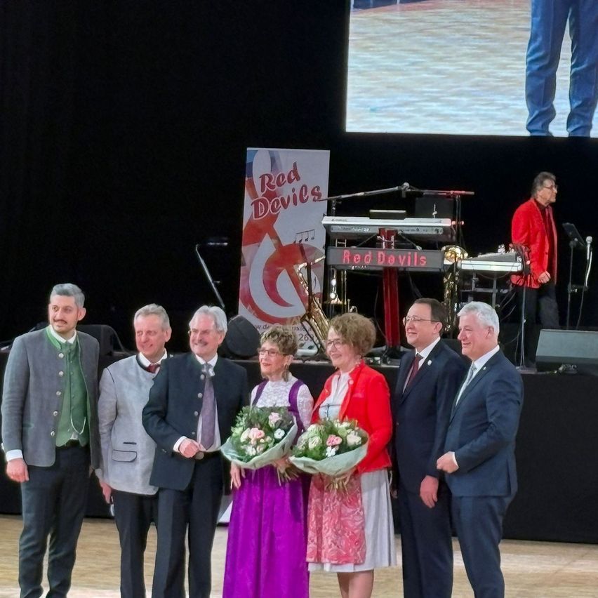 A group of people in formal attire stand on a stage, with a band and a large screen in the background. Two women hold flower bouquets.