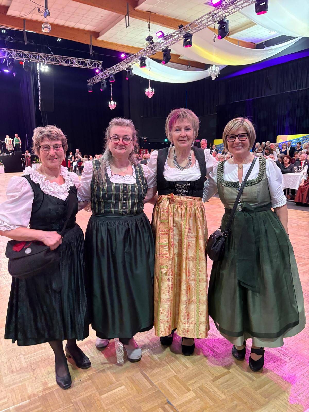 Four women in traditional attire are posing for a photo on a stage with a crowd in the background.