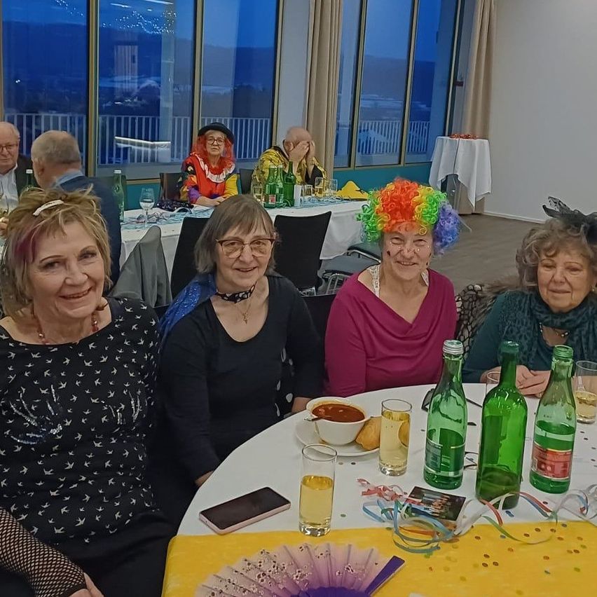 Mehrere Frauen und Männer sitzen um einen Tisch mit Essen, Getränken und einer festlichen Atmosphäre. Eine Frau trägt eine bunte Perücke. Sie feiern wahrscheinlich ein Ereignis.