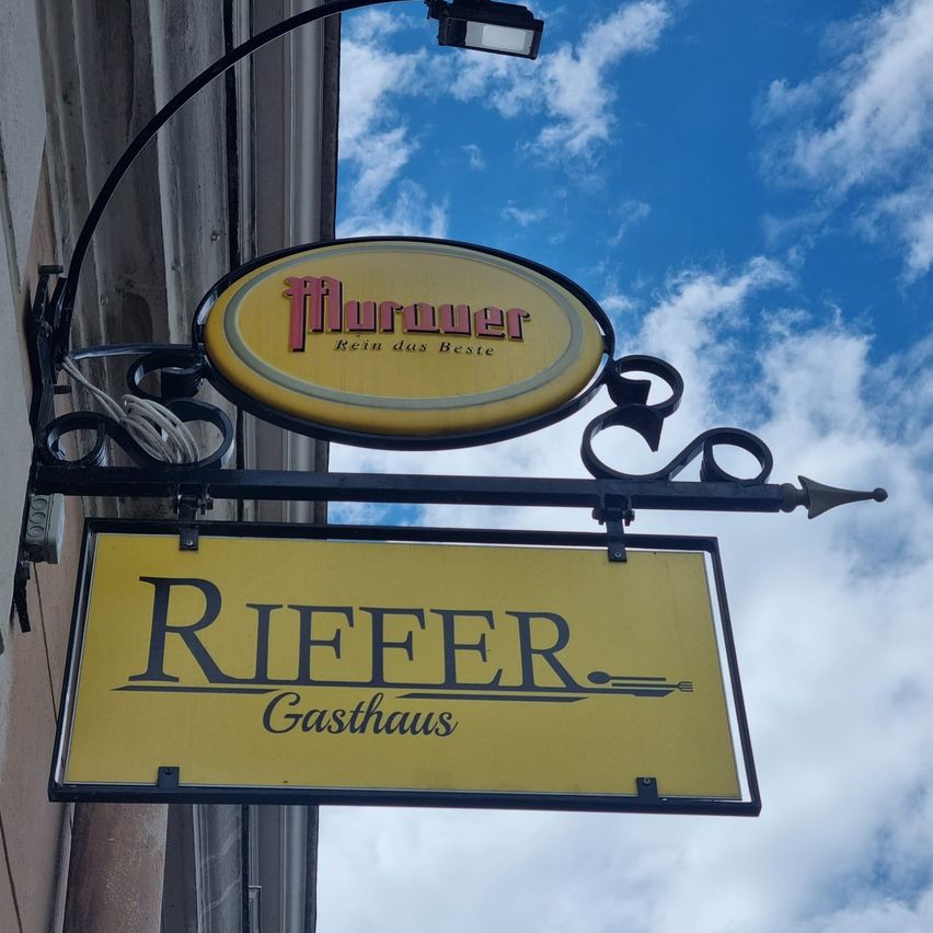 A yellow signboard for Riefer Gasthaus hangs from a metal bracket against a wall under a blue sky.