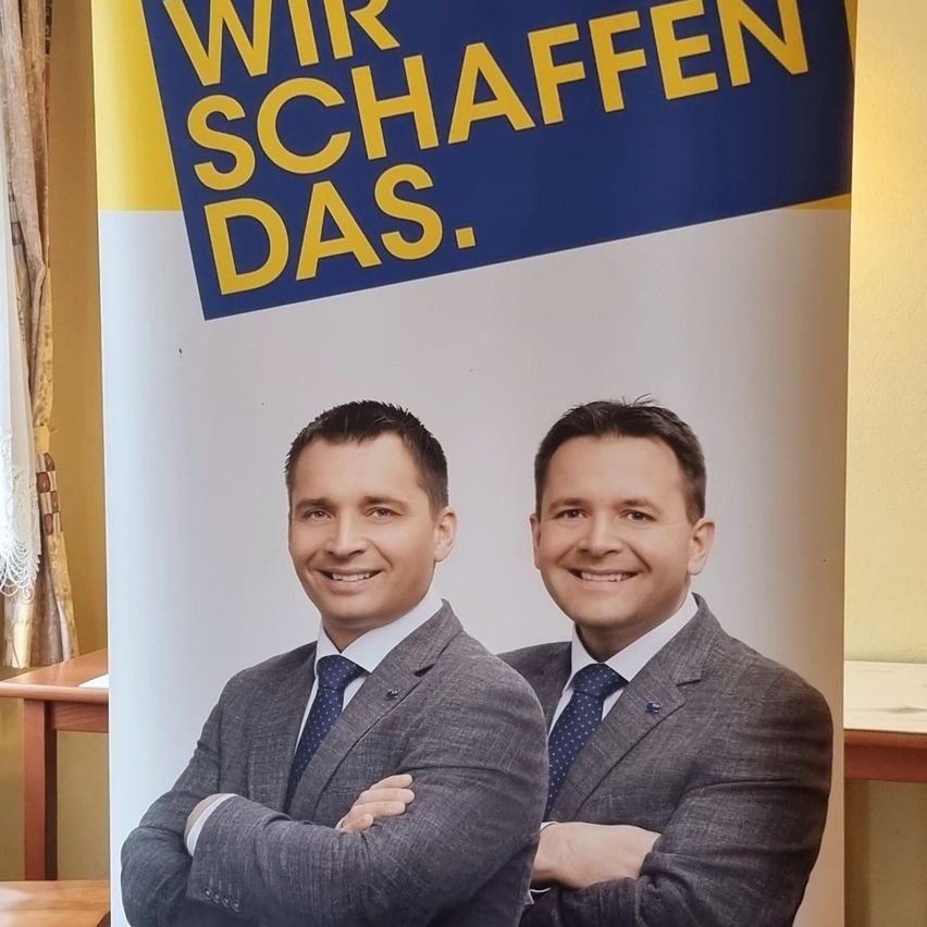 Two men are standing side by side, smiling, in a room with a blue and yellow banner above them. They both wear suits and ties.
