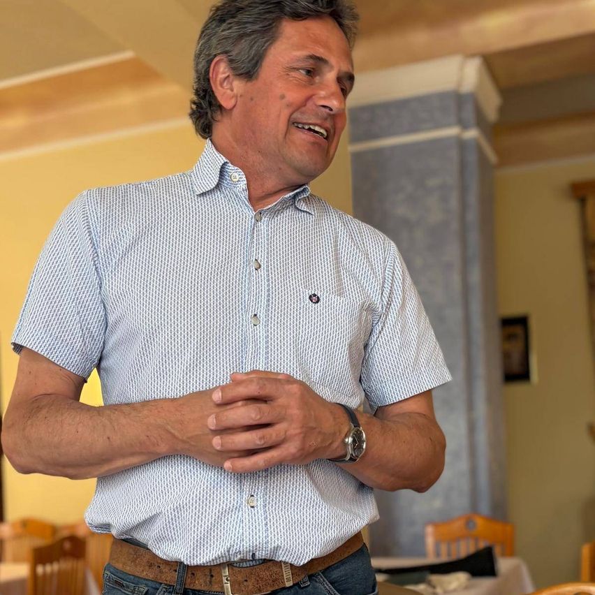 A man wearing a patterned shirt and a watch is standing in a room with his hands clasped together, smiling. Behind him, there are chairs and tables, and a picture frame is mounted on the wall.