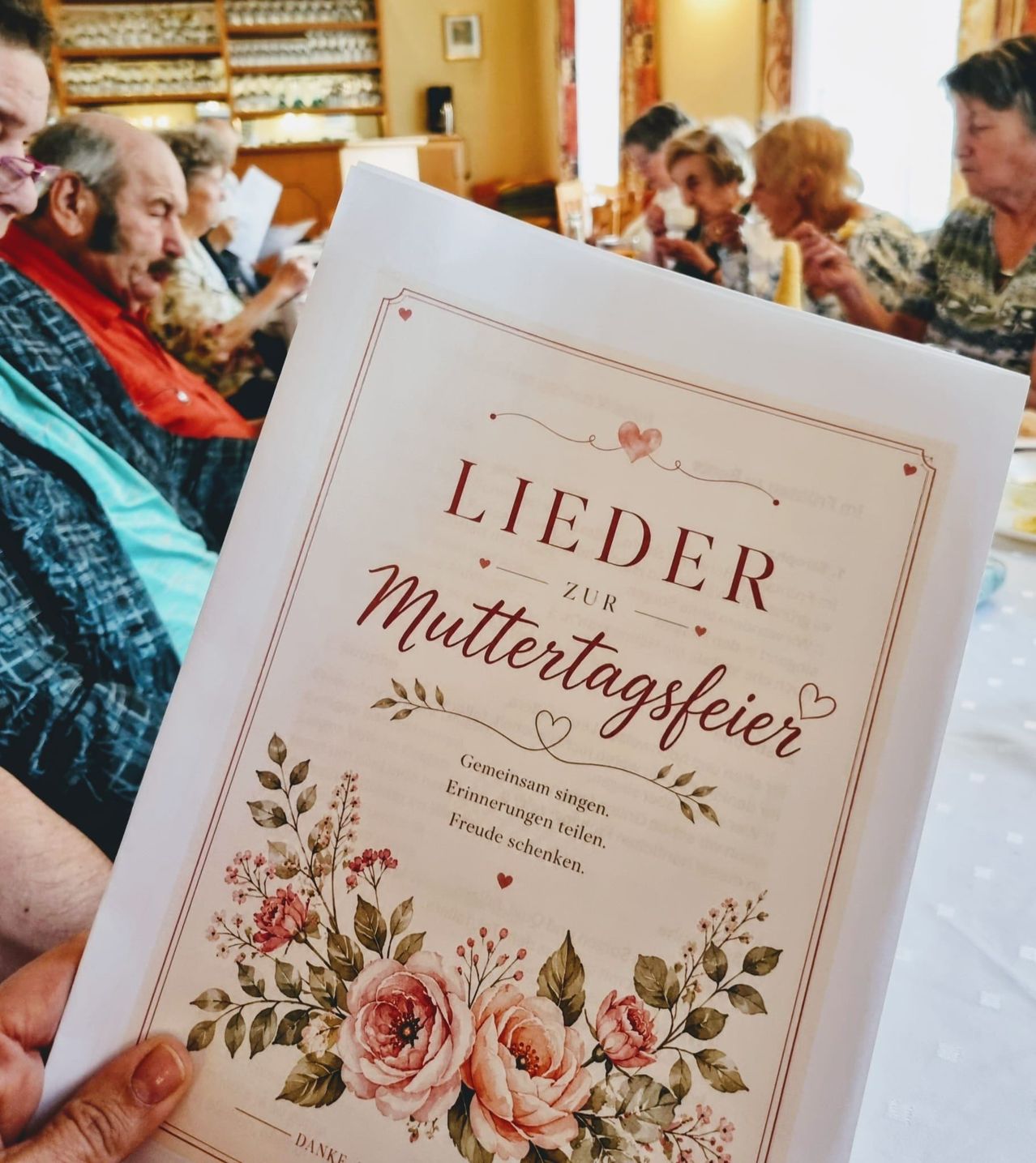 A person holding a booklet titled 'Lieder zur Muttertagsfeier' with a floral design and a group of people in the background.