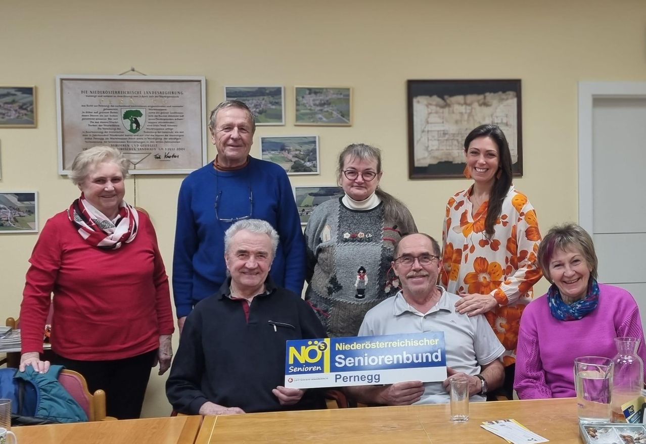 Gruppe von Senioren mit einem blauen Schild, auf dem 'NO Senioren' steht. Sie posieren für ein Foto in einem Raum mit gelben Wänden und gerahmten Bildern.