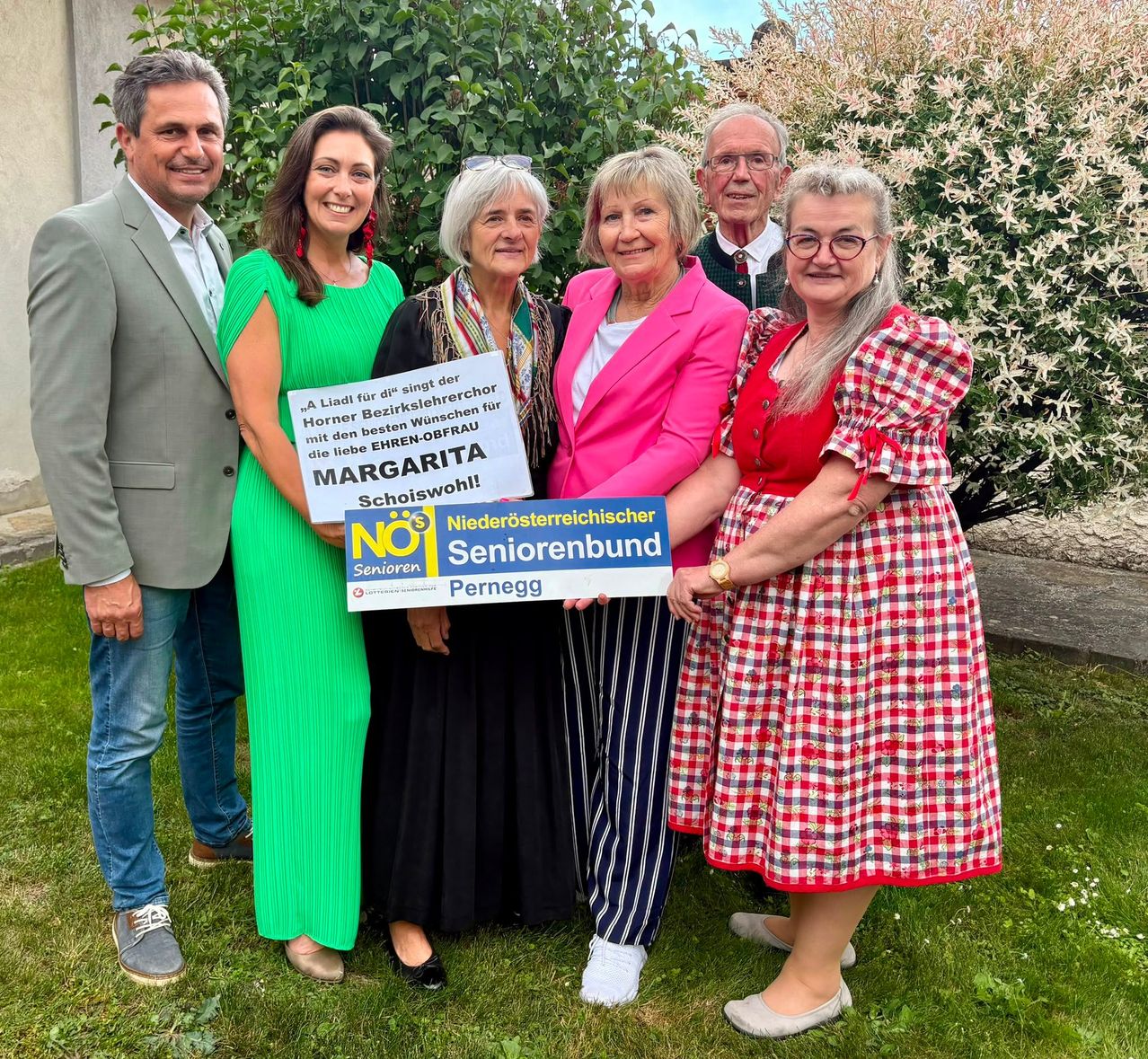 A group of people pose for a photo in front of a building with plants. They hold a sign with the text 'NO1 Seniorenbund Pernegg'.