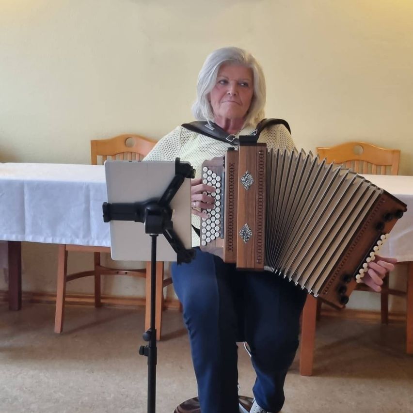An elderly woman with white hair sits in front of a table playing an accordion. Behind her, two wooden chairs and a table with a white tablecloth are visible.