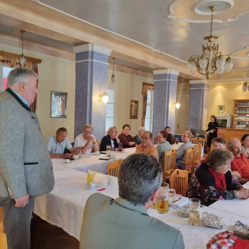 A group of adults sits at a table inside a dining room. The room has a chandelier and a cabinet with bottles. The man standing is looking at the people sitting.