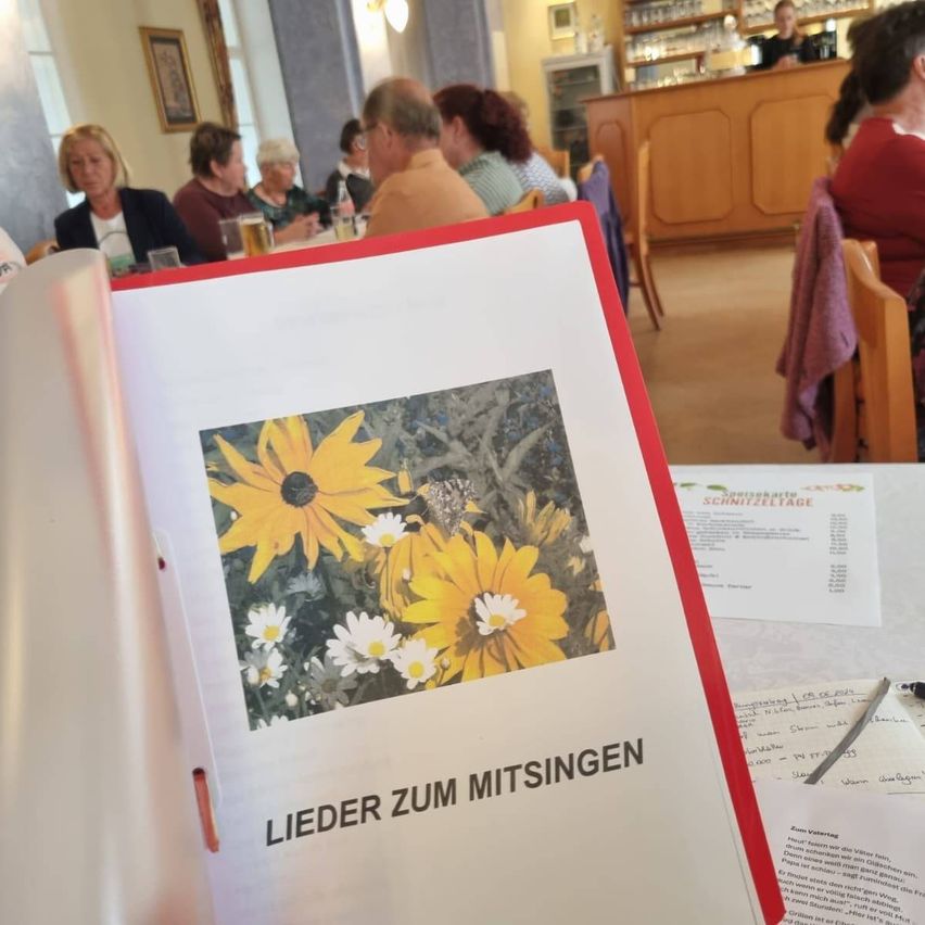 A photo of a group of people sitting at a table, with a booklet titled 'Lieder zum Mitsingen' open to a page with a floral image.