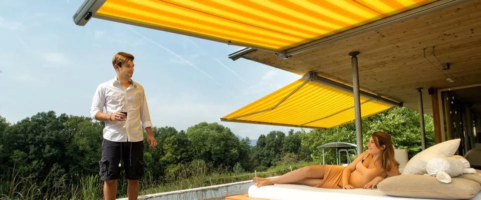 Ein Mann und eine Frau sind auf einer Terrasse. Der Mann steht und hält ein Getränk. Die Frau liegt auf einem Bett unter einem gelben Sonnensegel.