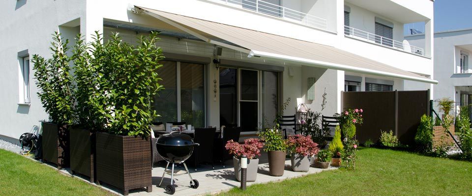 Ein Patio mit einem weißen Vordach und mehreren Topfpflanzen, darunter Rosen, und einem Grill auf der Seite. Ein weißes Haus mit Glasfenstern und einem Balkon im Hintergrund.