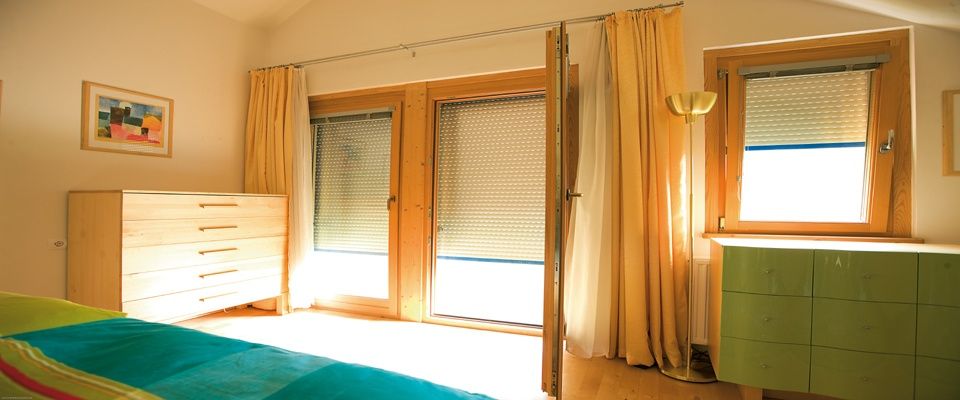 Ein Schlafzimmer mit Holzmöbeln, einem Bett, einer hölzernen Kommode und zwei großen Fenstern mit Jalousien und Vorhängen. Es gibt eine Stehlampe und einen grünen Schrank.