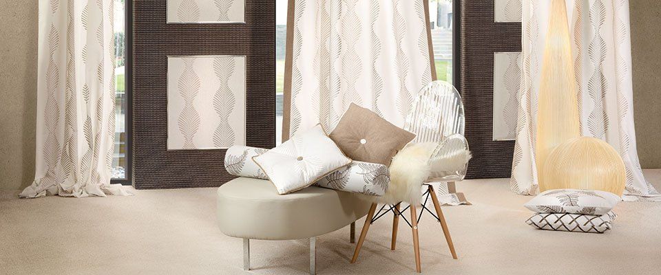 Ein Wohnzimmer mit einem beigefarbenen Ottoman, einem Stuhl mit Kissen und dekorativen Kissen, mit weißen Vorhängen und gemusterter Tapete.