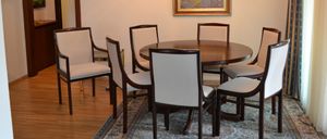 Ein runder Holztisch mit sechs Stühlen steht auf einem Teppich. Ein Bilderrahmen ist an der Wand hinter den Stühlen montiert. Ein Holzschrank steht links.