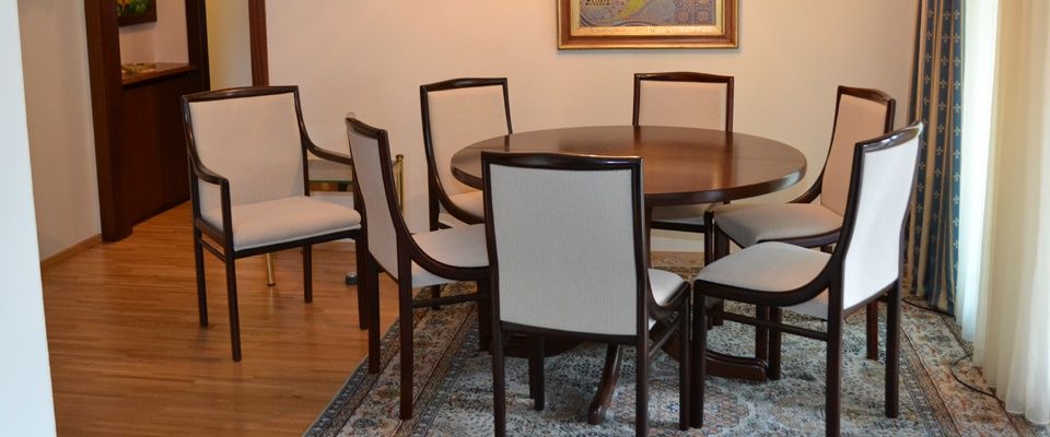 Ein runder Holztisch mit sechs Stühlen steht auf einem Teppich. Ein Bilderrahmen ist an der Wand hinter den Stühlen montiert. Ein Holzschrank steht links.