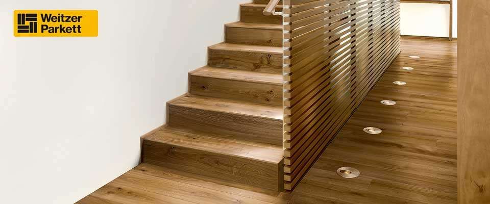 Ein Bild einer modernen Holztreppe mit einer weißen Wand, einem Handlauf und Bodenlampen. Der Text 'zeitzer rkett' erscheint in der oberen linken Ecke.