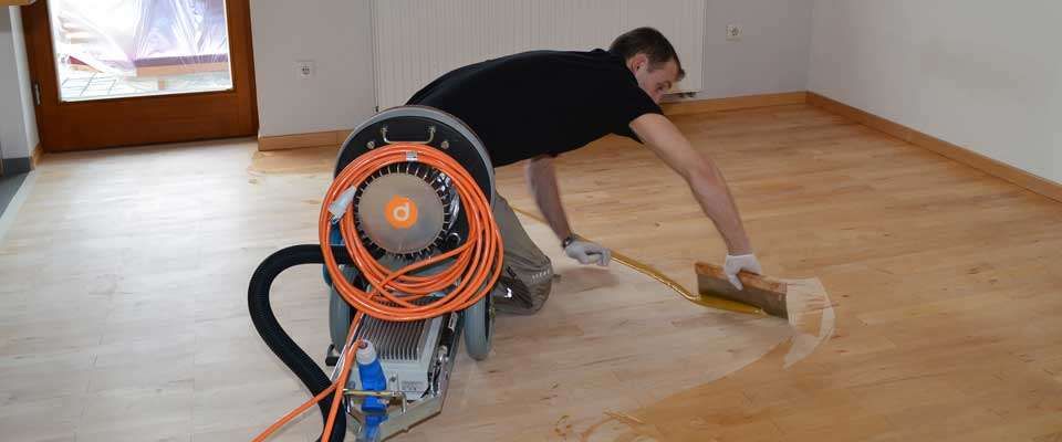 Ein Mann in einem schwarzen T-Shirt und weißen Handschuhen benutzt eine Fußbodenschleifmaschine zum Schleifen eines Holzbodens.