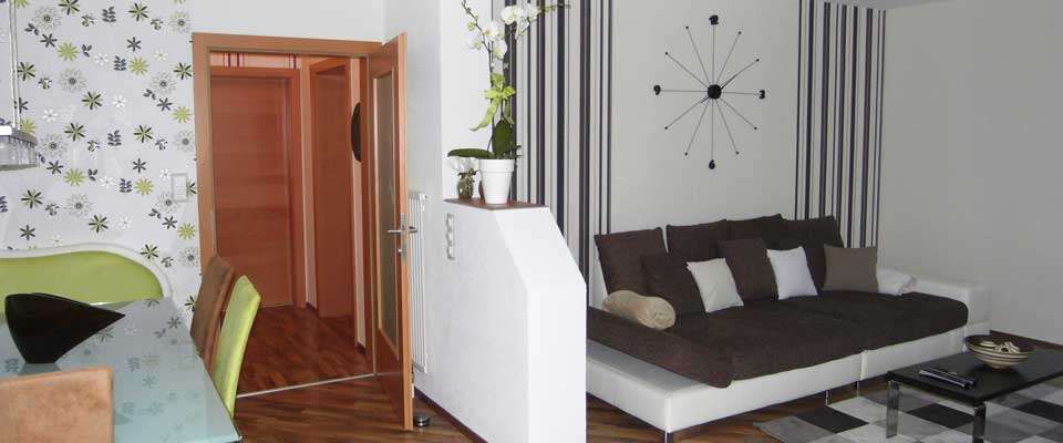 Ein leeres Wohnzimmer hat eine Couch, einen Heizkörper, eine Uhr und eine Topfpflanze auf einem weißen Regal. Ein Glastisch und Stühle befinden sich in der Nähe einer offenen Tür.