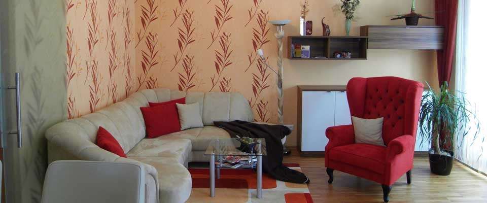 Ein Wohnzimmer mit einem beigen Sofa, roten Kissen und einem Glastisch. Der Raum hat einen roten Sessel, eine Lampe und Holzboden.