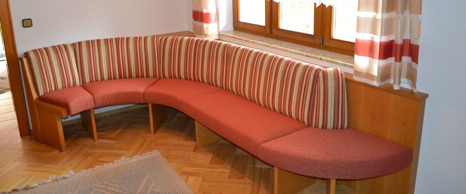 Eine gekurvte Bank mit roten Kissen und gestreiften Rückenlehnen steht in einer Ecke, nahe einem Fenster mit gestreiften Vorhängen. Der Boden ist aus Holz.