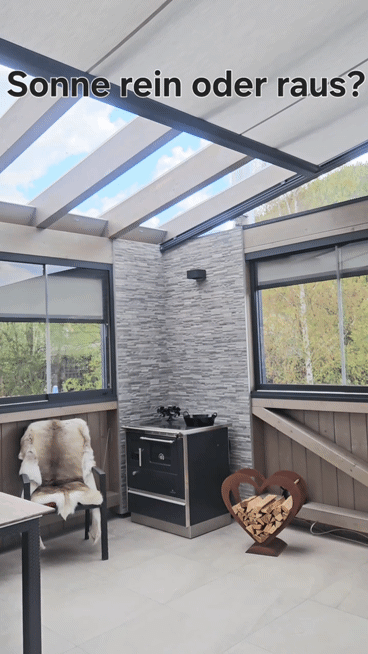 Ein Raum mit Glasdach und Wänden, ausgestattet mit einem Fellstuhl, einem Ofen und einem Holzkorb. Draußen sind Bäume durch die Fenster zu sehen.