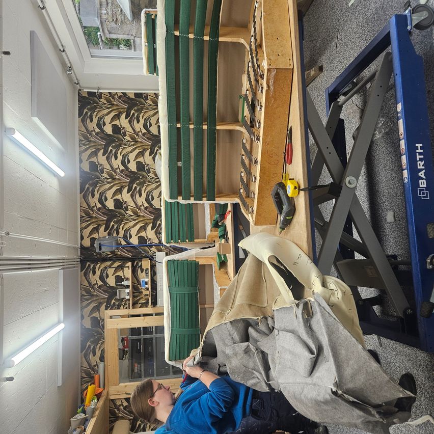Eine Frau in einem blauen Shirt arbeitet in einer Werkstatt mit einem großen Werktisch. Verschiedene Werkzeuge und Materialien sind verstreut, einschließlich eines Stoffstücks auf einem blauen Wagenheber. Die Werkstatt verfügt über eine gemusterte Wand und Deckenleuchten.