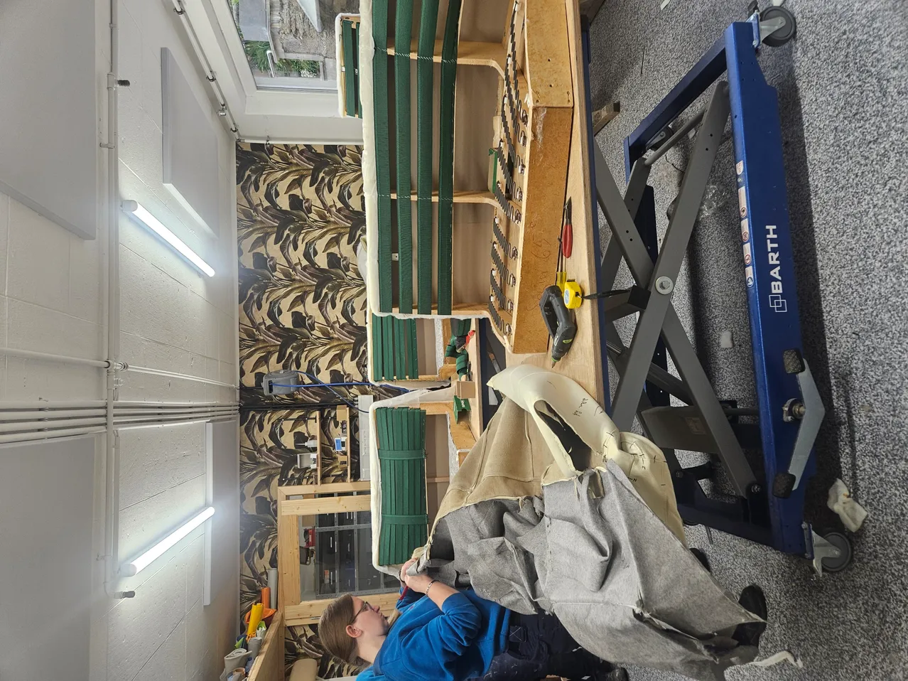 Eine Frau in einem blauen Shirt arbeitet in einer Werkstatt mit einem großen Werktisch. Verschiedene Werkzeuge und Materialien sind verstreut, einschließlich eines Stoffstücks auf einem blauen Wagenheber. Die Werkstatt verfügt über eine gemusterte Wand und Deckenleuchten.