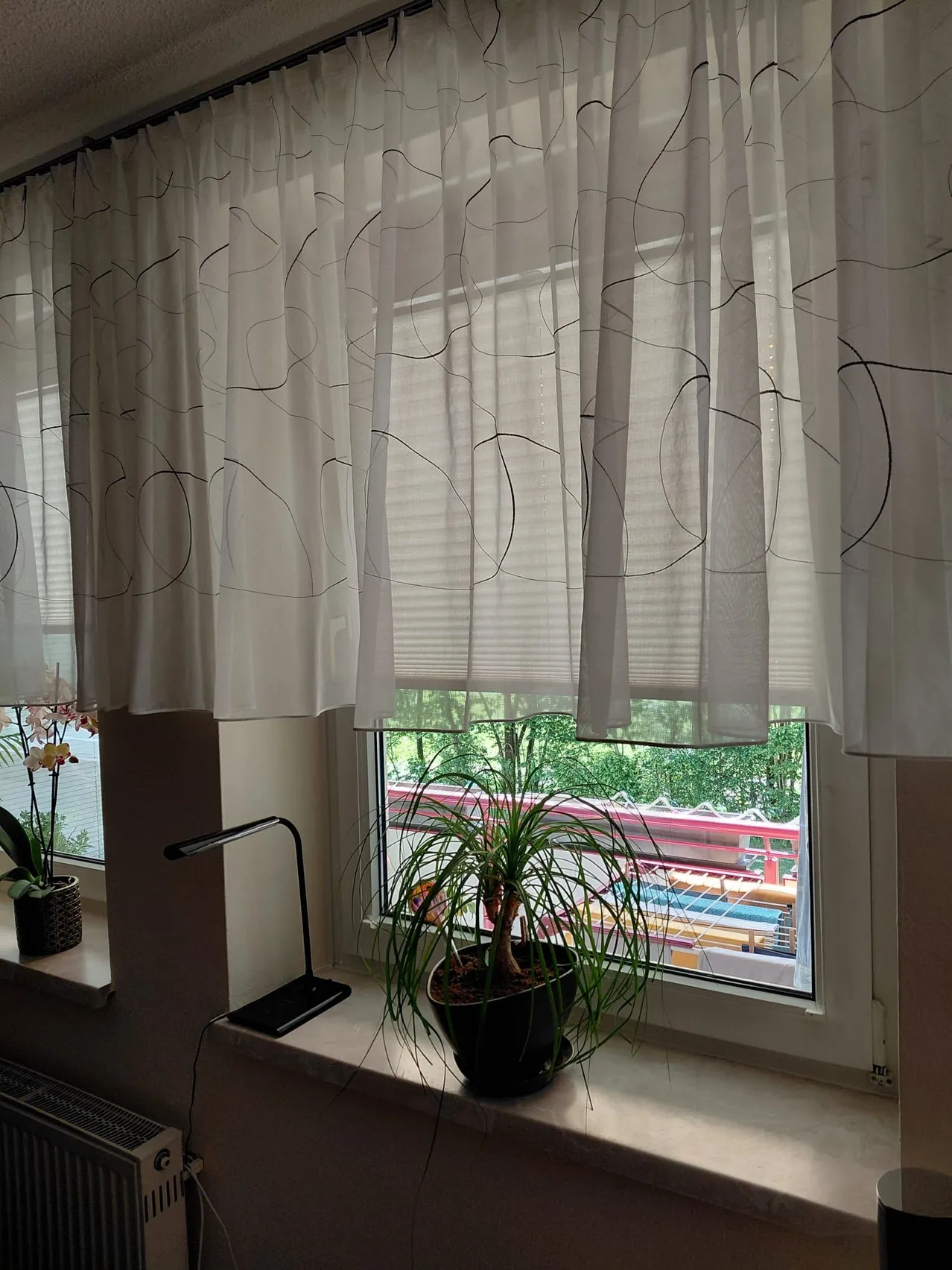Ein Fenster mit weißen Vorhängen und einem gemusterten Design, einer Lampe und einem Topf auf der Fensterbank, mit Blick auf einen Zug auf der Schiene.