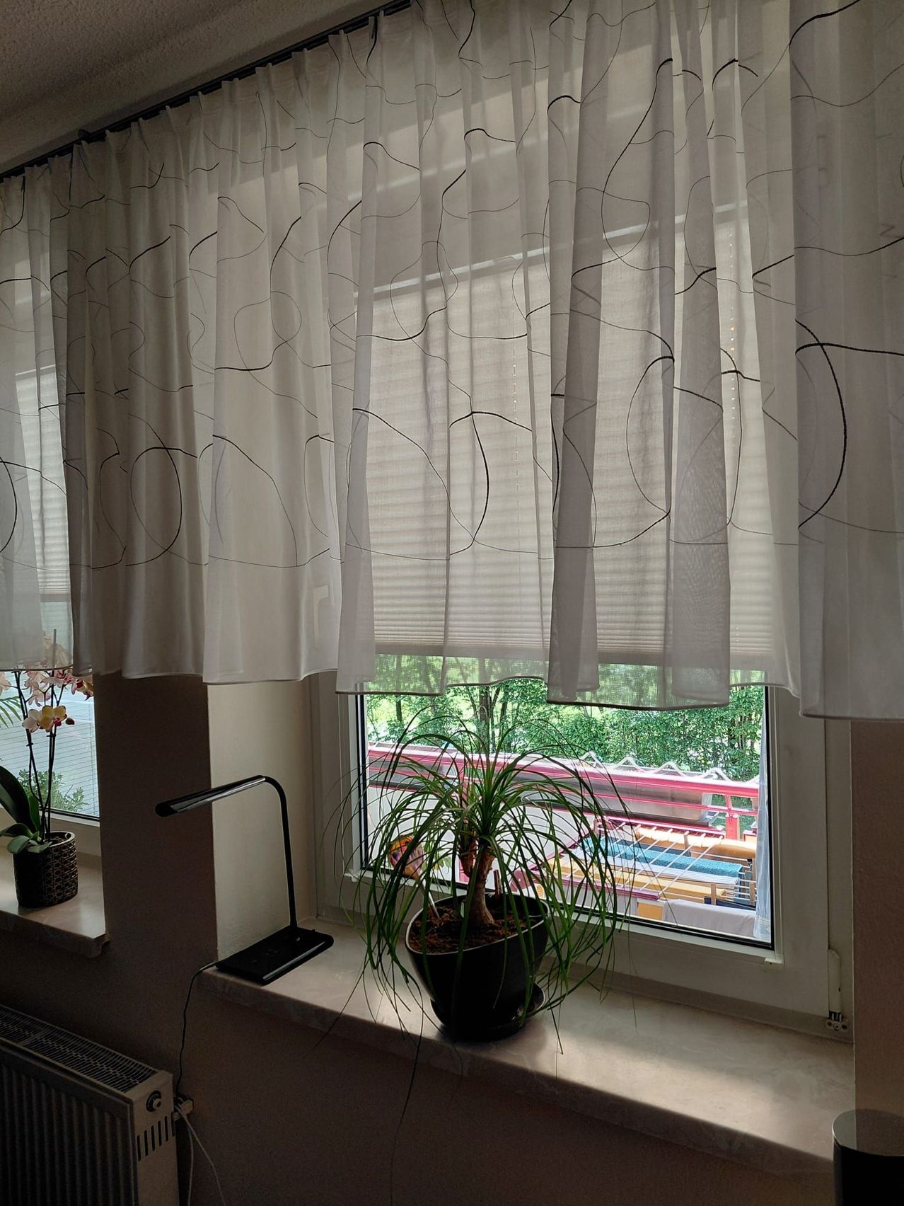 Ein Fenster mit weißen Vorhängen und einem gemusterten Design, einer Lampe und einem Topf auf der Fensterbank, mit Blick auf einen Zug auf der Schiene.