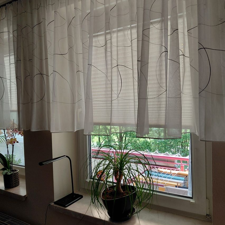 Ein Fenster mit weißen Vorhängen und einem gemusterten Design, einer Lampe und einem Topf auf der Fensterbank, mit Blick auf einen Zug auf der Schiene.