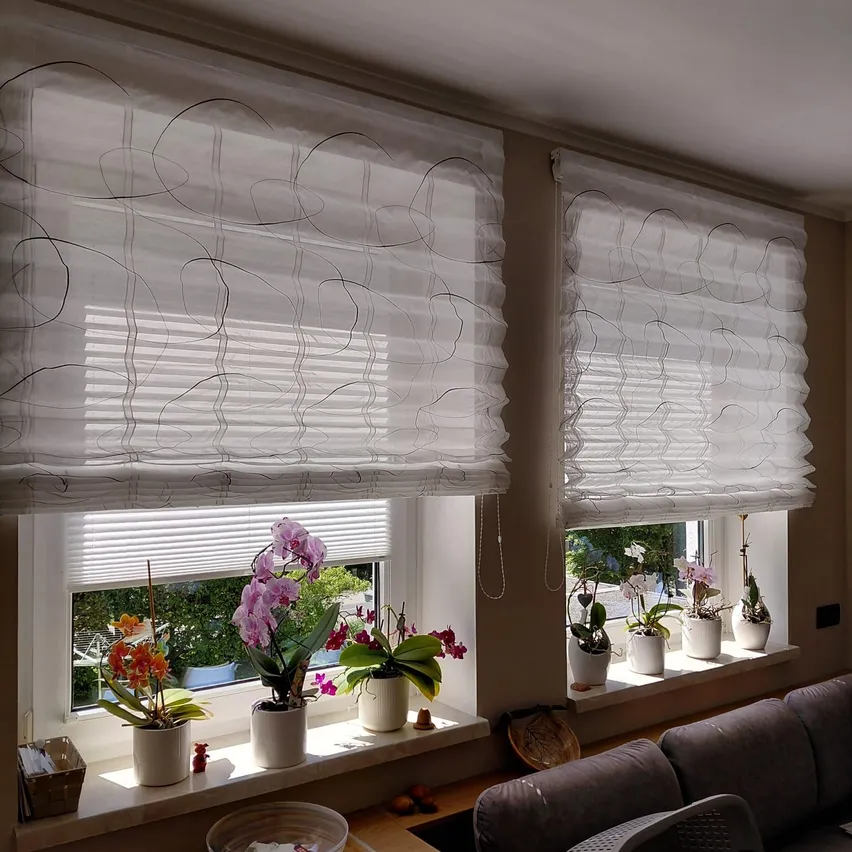 Zwei Fenster mit weißen gemusterten Vorhängen und weißen Jalousien, mit Topfpflanzen auf den Fensterbänken und einem Sofa davor.