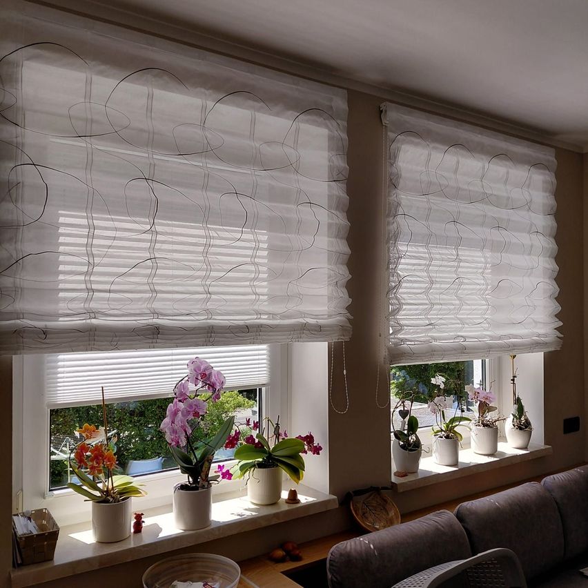 Zwei Fenster mit weißen gemusterten Vorhängen und weißen Jalousien, mit Topfpflanzen auf den Fensterbänken und einem Sofa davor.