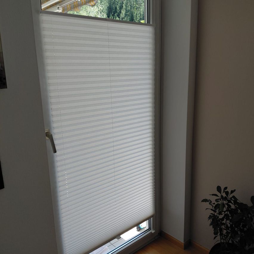 Ein weißer vertikaler Rollladen ist über einem Fenster gezogen. Ein Topf mit Pflanzen steht neben dem Fenster. Eine weiße Wand ist rechts, und ein Fenster ist über dem Rollladen.