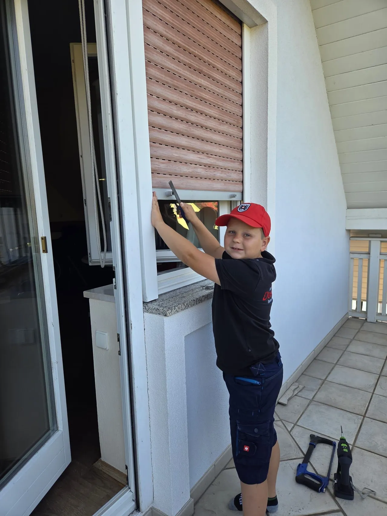 Ein junger Junge in einer roten Kappe und einem schwarzen Shirt repariert ein Fenster auf einem Balkon und hält ein Werkzeug. Der Balkon hat geflieste Böden und weiße Geländer.