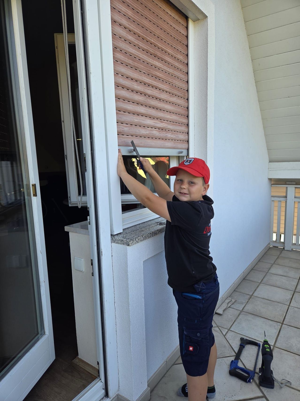 Ein junger Junge in einer roten Kappe und einem schwarzen Shirt repariert ein Fenster auf einem Balkon und hält ein Werkzeug. Der Balkon hat geflieste Böden und weiße Geländer.